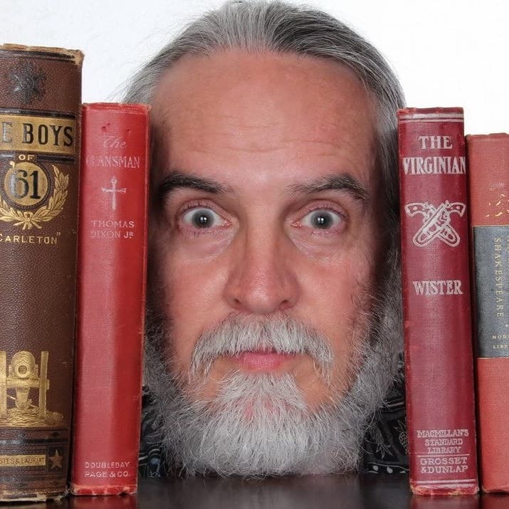 A wide-eyed man with a grey beard peers from between stacks of vintage books.